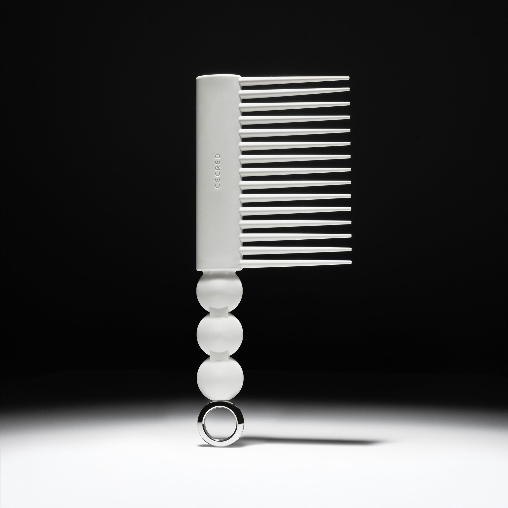 Cécred Rake Comb standing upright against a dark background with dramatic spotlighting, showcasing its wide tooth spacing, beaded handle, and circular ring